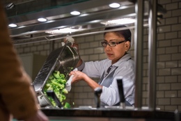 Mitarbeiterin an der Essensausgabe füllt frisches Gemüse in die Auslage