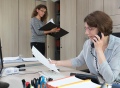 Vorschau: Zwei Mitarbeiterinnen im Büro, eine am Schreibtisch sitzend in einem telefonischen Gespräch, die andere stehend mit Aktenordner am Schrank