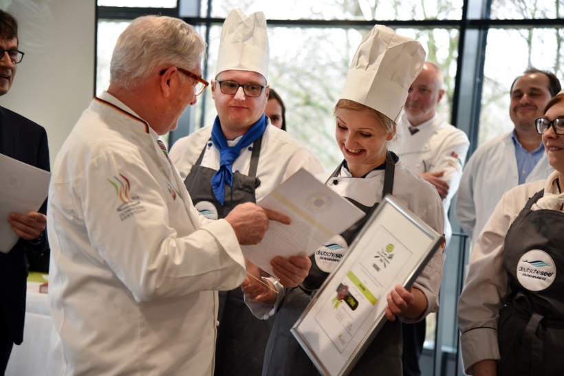 Jürgen Rabe, Prüfer und 2. Vorsitzender des Verbandes der Köche Deutschlands | Landesverband Nordrhein-Westfalen, überreicht Kaya Grube eine Überraschungsprämie – eine Einladung zum Probetraining der Deutschen Nationalmannschaft der Köche.